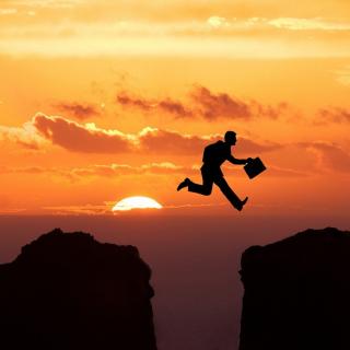 silhouette against a sunset of someone carrying a briefcase jumping across a deep crevice