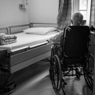 Elderly man, alone in a hospital room, sitting in a wheelchair next to an unmade hospital bed